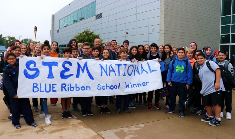 STEM students stand outside the school with a handmade banner reading STEM National Blue Ribbon School.