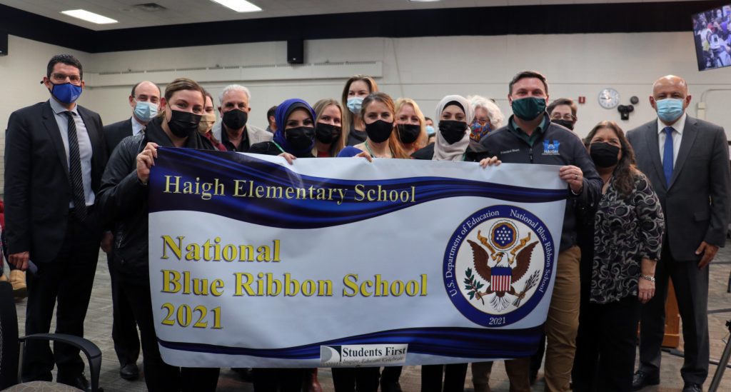 Posing with a sign celebrating Haigh Elementary receiving a 2021 National Blue Ribbon School Award is a group of people including some school staff, the principal, Board of Education members and Superintendent Glenn Maleyko.