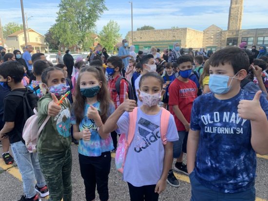 A group of elementary students outside in face masks give a thumbs up sign.