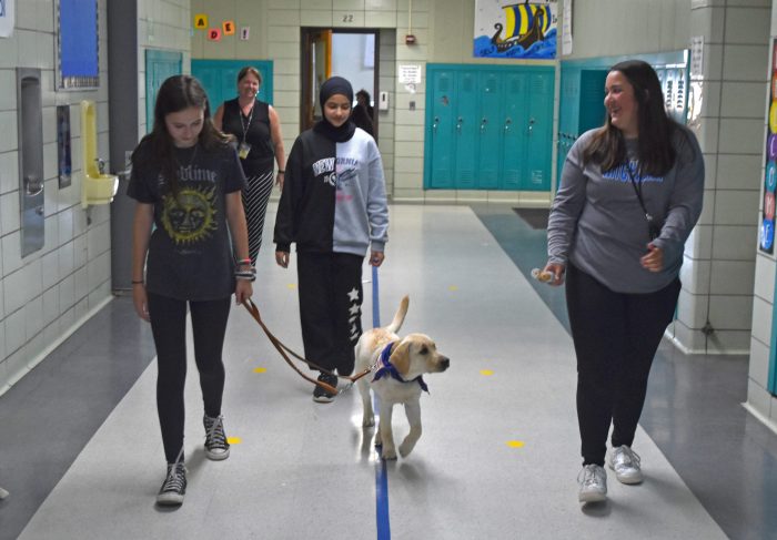 Three girls, followed by a teacher, walk a young dog down a school hall.