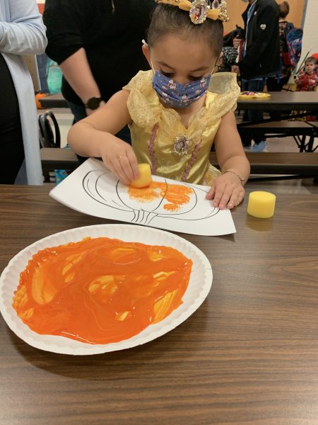 A child uses a sponge and a plate of orange paint to color a pumpkin picture.