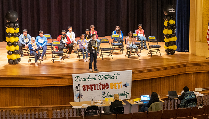 A group of students sits on the stage while one student spells a word during the 2022 Dearborn Public Schools Spelling Bee.