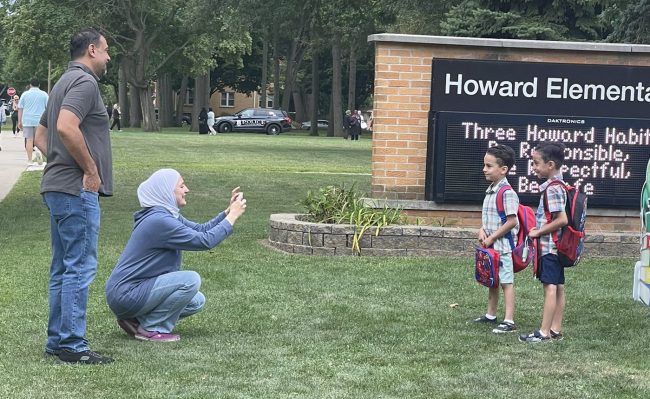 A mother snaps a photo and a father watches as two young students pose in front of the Howard Elementary School sign on the first day of school in August 2022.