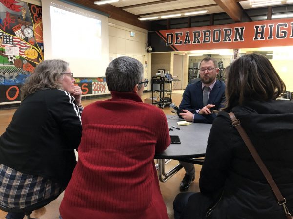 Four people sit and talk at a table in the Dearborn High School cafeteria during a town hall meeting.