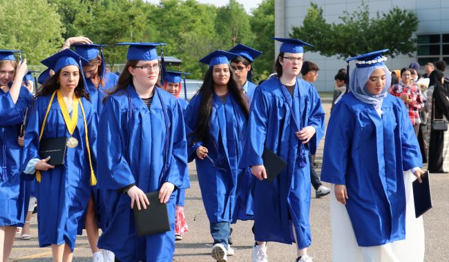 Several Virtual K-12 graduates in blue caps and gowns walk across a parking lot