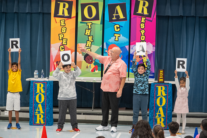 Four students with signs spelling out ROAR stand with Al the Respect Guy during his show at Geer Park Elementary School in September 2023.