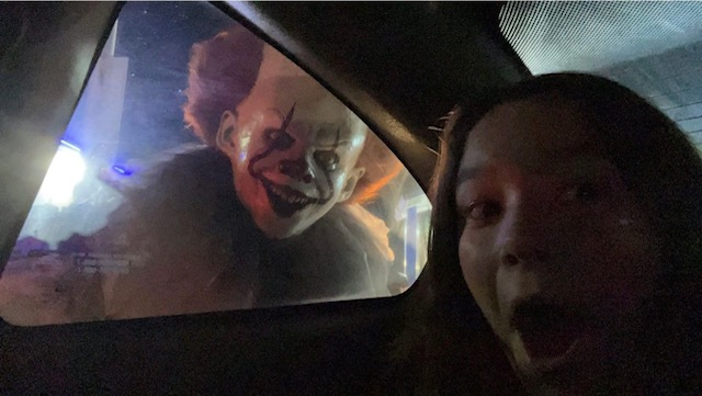 A spooky clown peers in a car window as a girl inside the car yells.