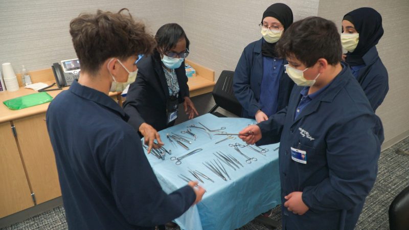 Four Henry Ford Early College students work with a woman learning about different surgical instruments and how to handle them.