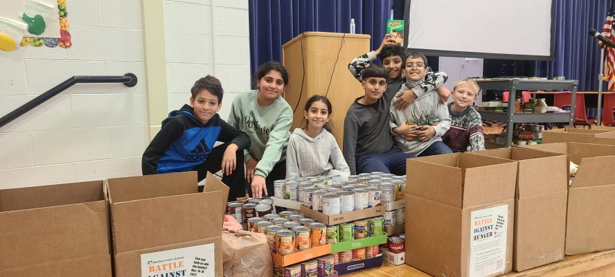 Six children sit on a school stage surrounded by boxes and trays of canned food.