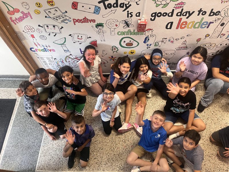 About a dozen students sit in a hallway near a partially completed mural that says "Today is a good day to be a leader."