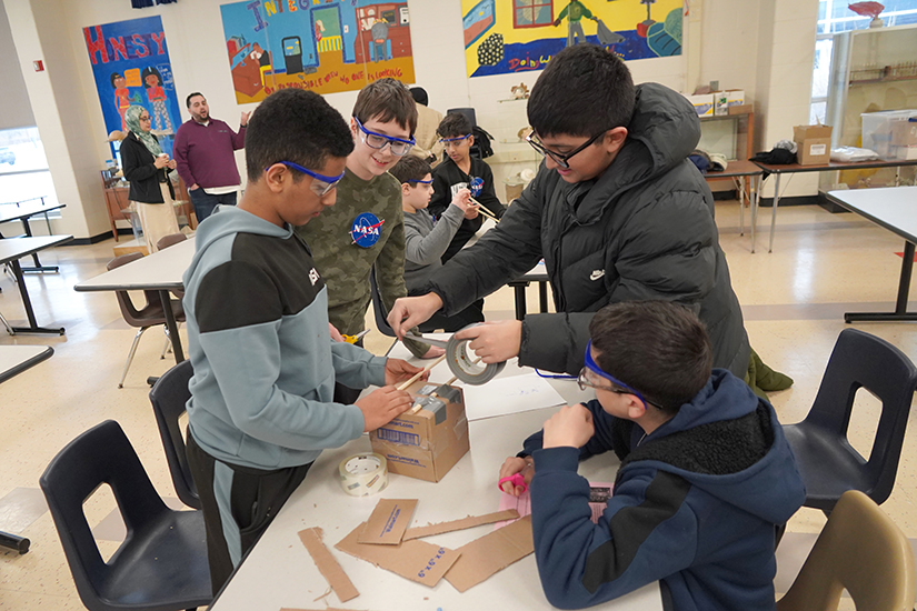 Four boys work on a cardboard crane, with two applying duct tape.