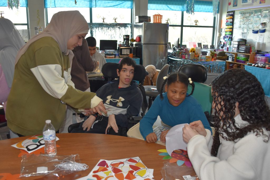 Volunteers help two students in wheelchairs decorate turnkey masks.