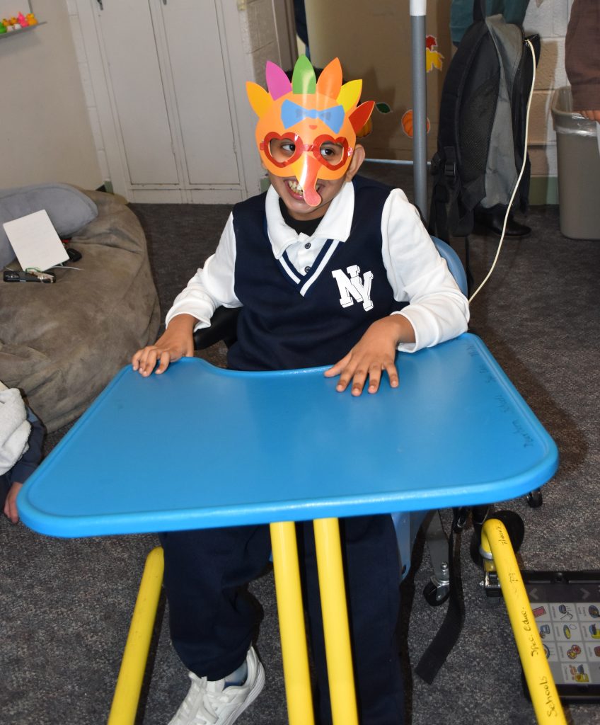 A student in a wheelchair smiles wearing his paper turkey mask.