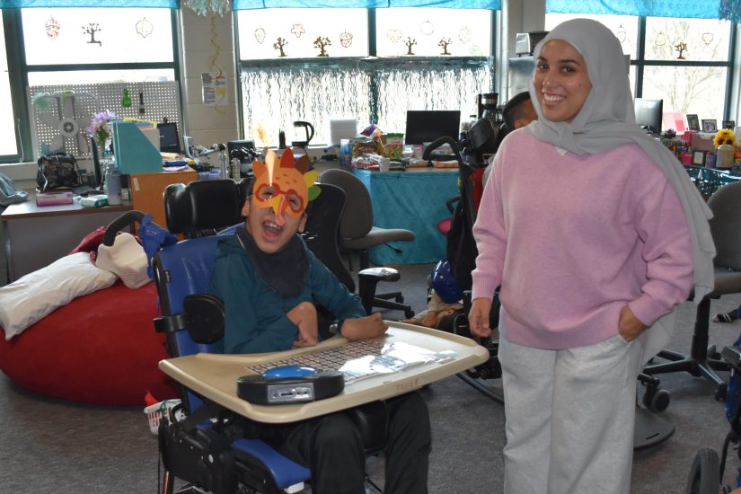 A student in a wheelchair smiles wearing a paper turkey mask a volunteer standing nearby helped him make.