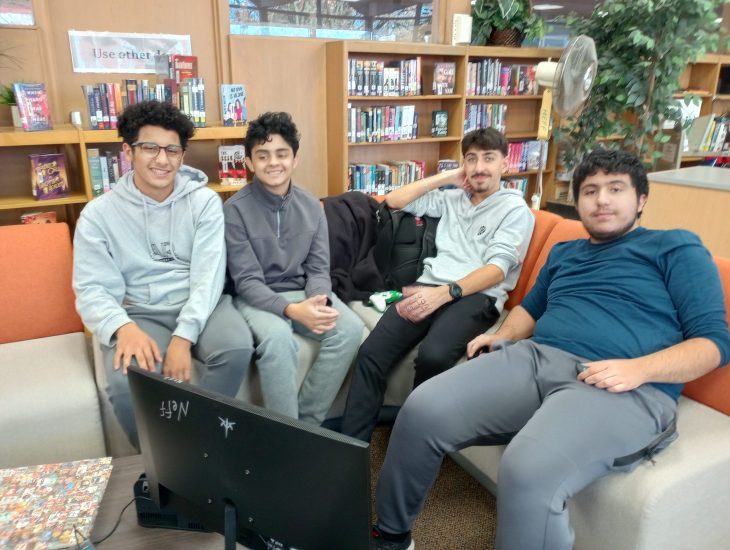 Four students sit on a library couch in front of a television.