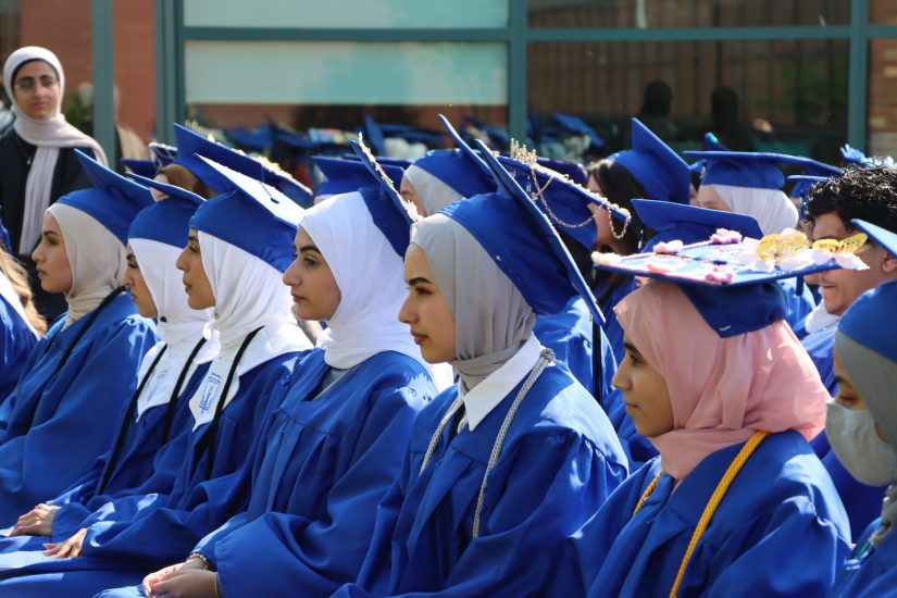 Students in blue caps and gowns sit outside during a graduation ceremony.