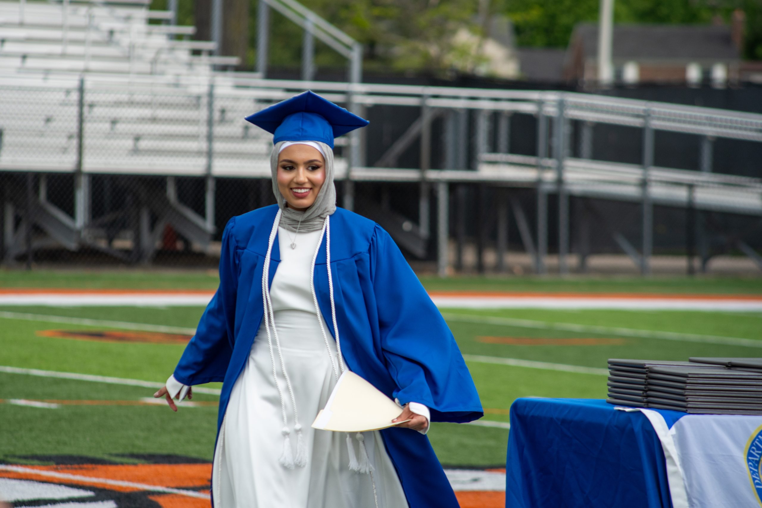 A girl in a blue cap and gown walks across a football field